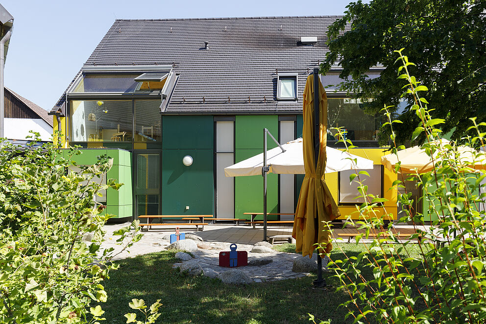 Blick an Sträuchern vorbei in den Garten mit Wasserlauf, das Kinderhaus im Hintergrund